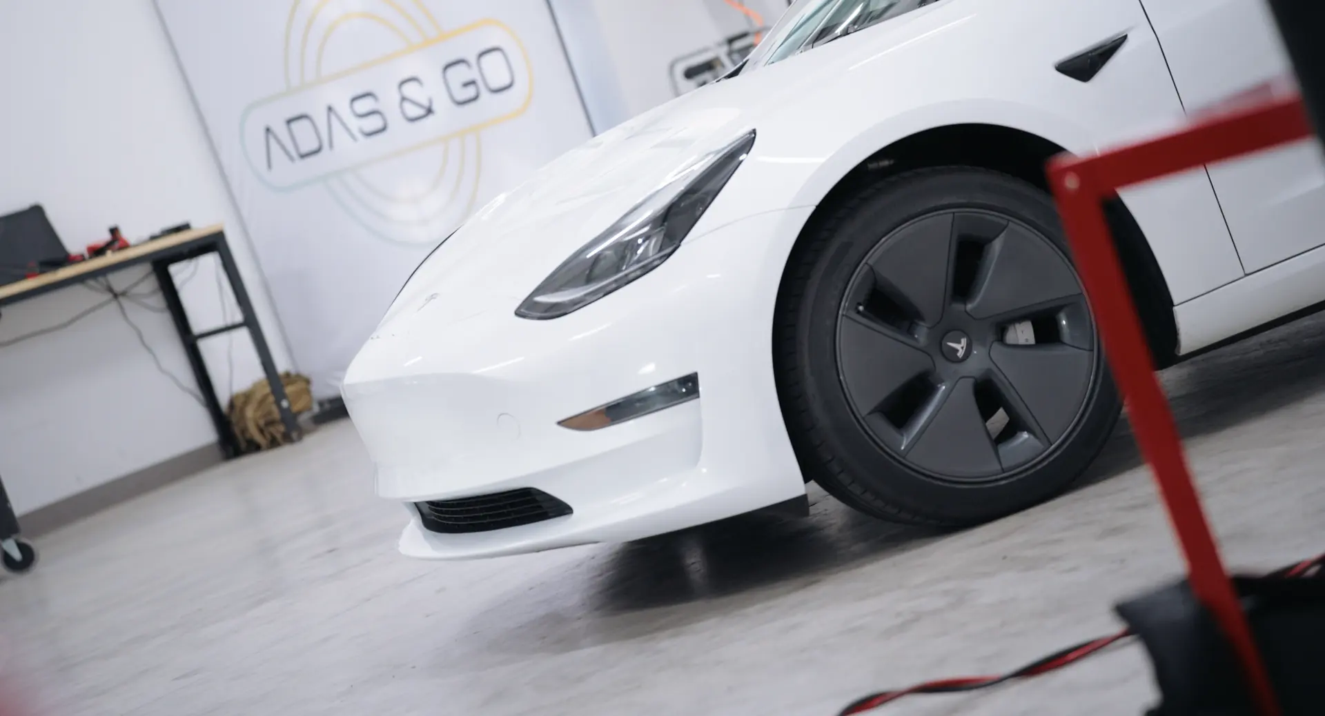 A white electric sedan on the alignment rack in front of the ADAS & GO wall logo.