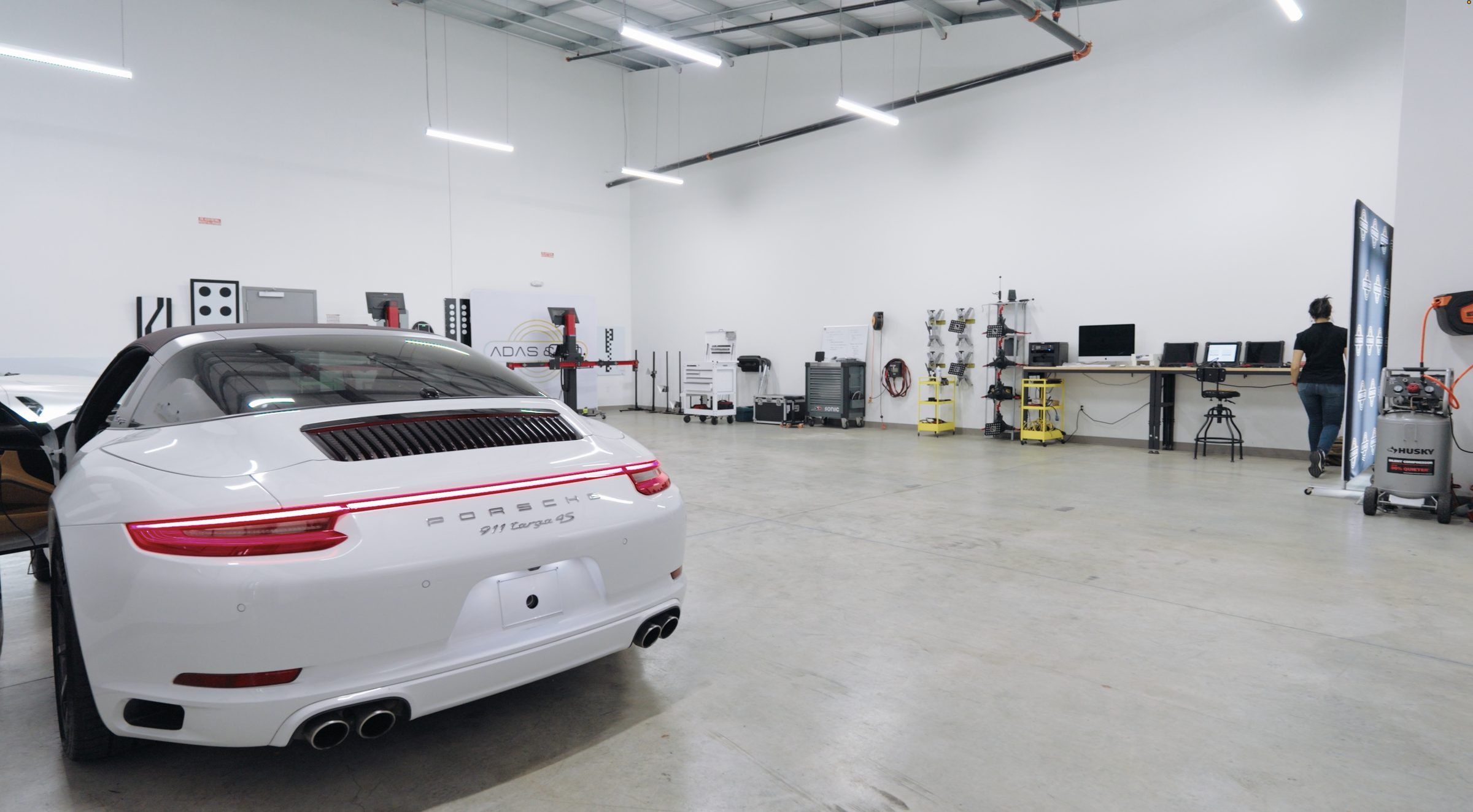 A white Porsche parked in an ADAS & GO static calibration laboratory, with equipment bays and technician workstations along the back wall.
