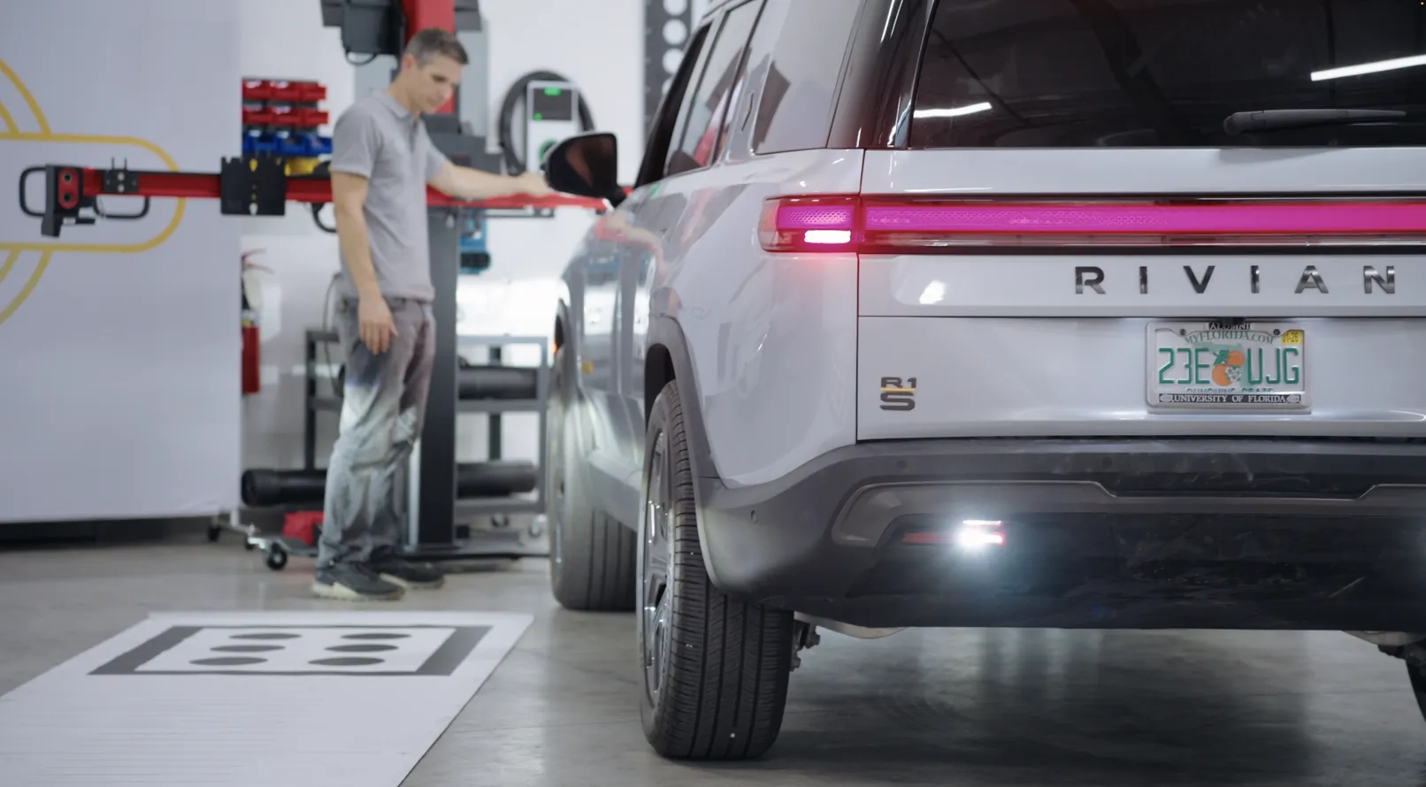 A technician holding a calibration target bar behind a white Rivian SUV standing on a printed floor target.