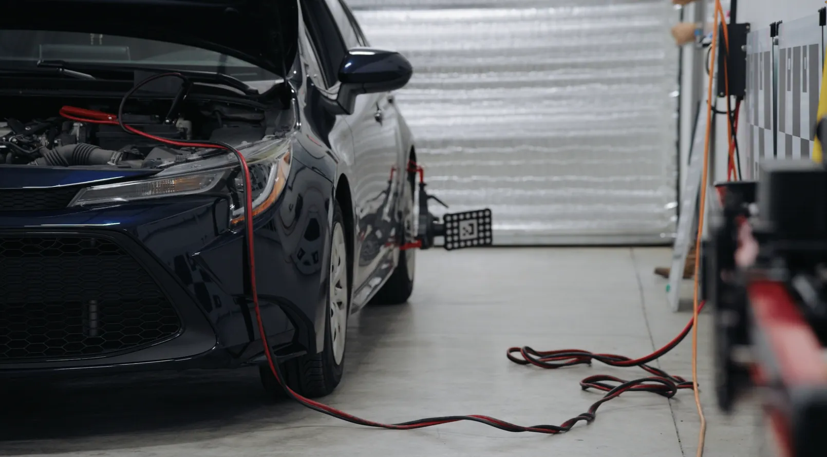 A sedan with its hood open and charging leads connected, parked in a service bay.