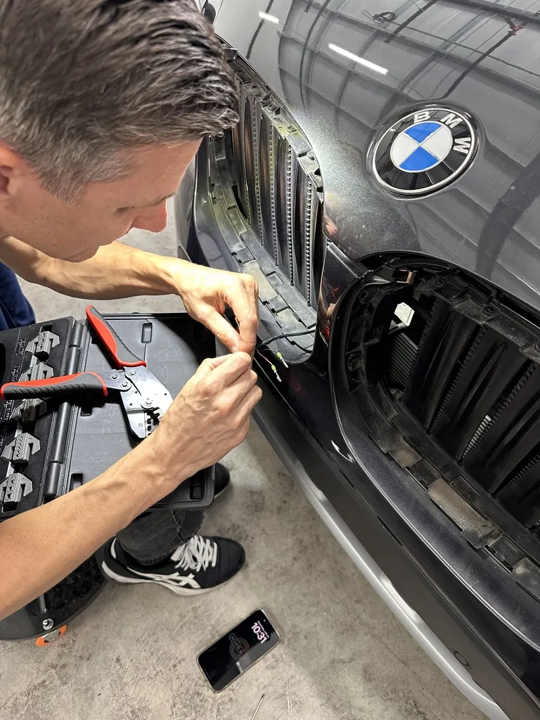 A technician reaching into the front of a BMW to work on the wiring behind the grille.